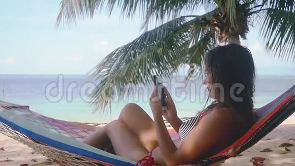 穿比基尼的漂亮年轻黑发女人在靠近大海和棕榈树的热带海滩的吊床上使用手机慢慢视频的预览图