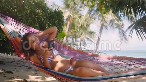 漂亮的比基尼女郎使用移动电话在海边海滩的吊床上聊天和棕榈树慢慢视频的预览图