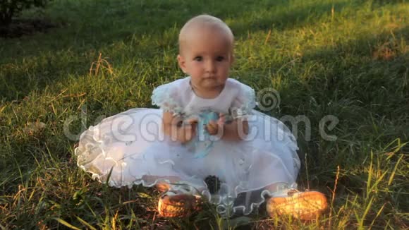 一个小女孩穿着白色连衣裙坐在草地上看着相机然后看着远处夏天日落视频的预览图