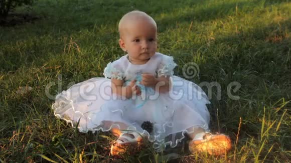 一个小女孩穿着白色的裙子坐在草地上看着镜头把草叶放进嘴里视频的预览图