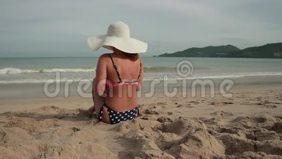 年轻女子在海滩上晒日光浴时用手机穿着比基尼和太阳帽的漂亮女孩拍照视频的预览图