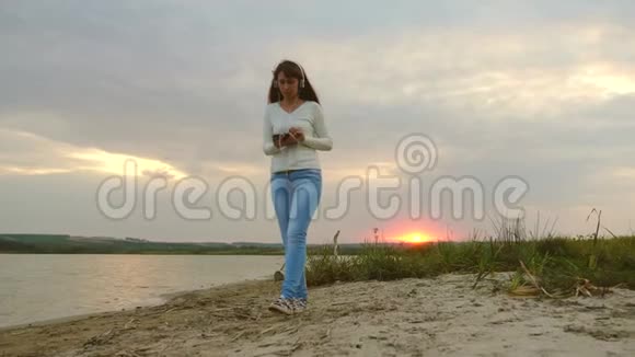 阳光下的女孩在沙滩上散步在平板电脑上查看邮件带着耳机的女孩走在海滩上视频的预览图