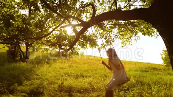 快乐的女孩留着长发在树枝上荡秋千在金色夕阳的明亮光线下微笑慢动作视频的预览图