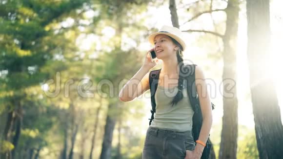 年轻的旅游女孩戴着帽子在夏天的森林里散步时与智能手机聊天视频的预览图