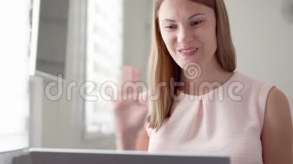 年轻女子坐在家里拿着笔记本电脑使用电脑聊天通过信使应用程序微笑着挥手致意视频的预览图