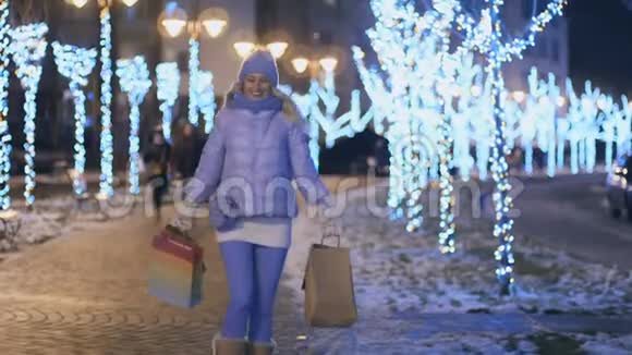 快乐的金发女郎带着包裹走在灯光之夜的城市冬天视频的预览图