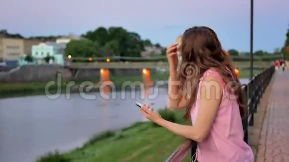 年轻漂亮的女孩使用智能手机带着耳机晚上在海滨老城区河边和视频的预览图