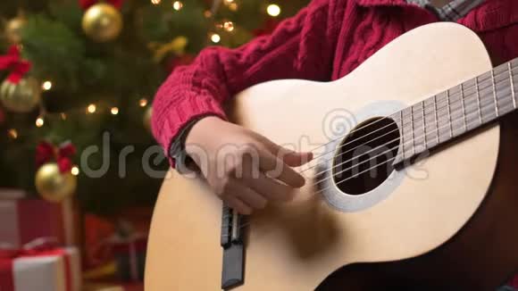 少年男孩弹吉他坐在室内靠近装饰好的圣诞树灯光打扮成圣诞老人助手圣诞快乐快乐视频的预览图