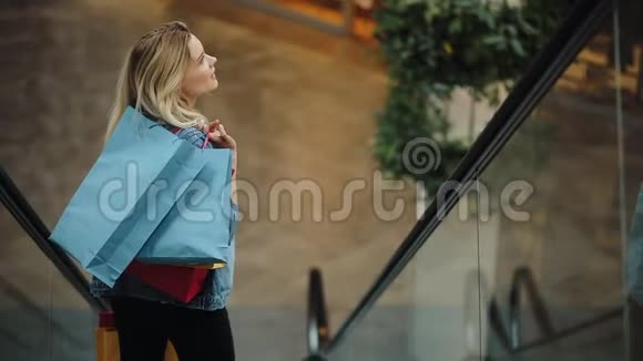 穿着牛仔裤夹克的金发女郎微笑着走下购物中心的移动楼梯视频的预览图