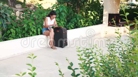 年轻的晒黑的黑发女人穿着短裤太阳镜和手提箱坐着在夏天使用智能手机视频的预览图
