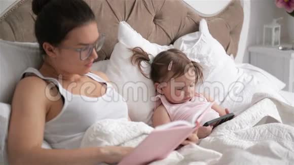 家庭日在卧室休息聪明的宝贝女孩正在使用智能手机戴眼镜的布鲁内特女士正在读一本书视频的预览图