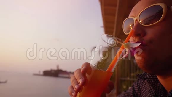 年轻的帅哥戴着太阳镜喝着鸡尾酒在海滩咖啡馆装饰花在令人惊叹的日落慢动作视频的预览图