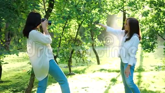 一位穿着白色衬衫的漂亮女孩职业摄影师正在拍摄一个快乐的微笑女孩在公园里的照片视频的预览图