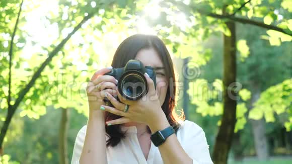 一位身穿白衬衫笑容可掬的女摄影师正在公园里柔和的背景下拍摄照片视频的预览图