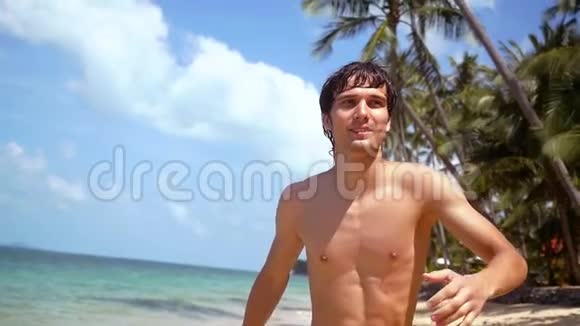 阳光明媚的一天年轻迷人的快乐男人在海滩上奔跑棕榈树和背景上的蓝天慢慢慢慢视频的预览图