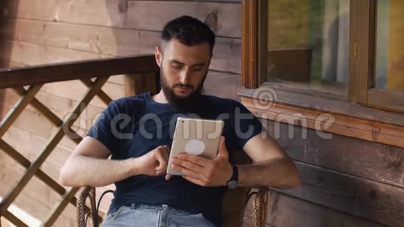 一个留胡子的年轻人坐在乡间别墅的阳台上用平板电脑视频的预览图