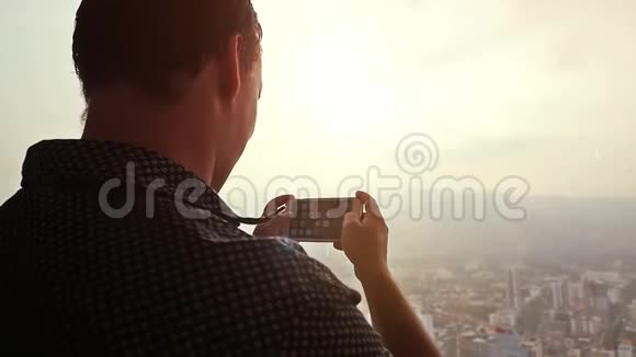 年轻的商人在令人惊叹的日落和美丽的城市景色下用手机在窗户旁边拍照慢动作视频的预览图