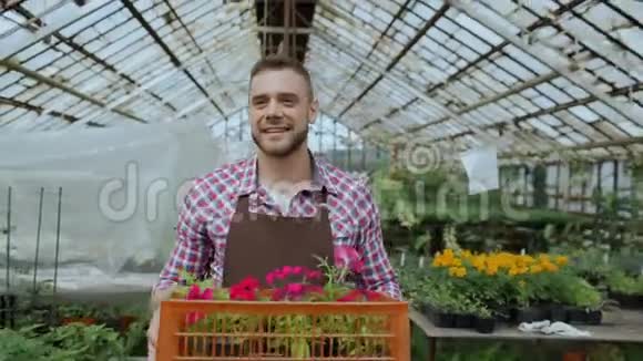 戴着围裙的快乐微笑的男人在温室里捧着鲜花的盒子视频的预览图