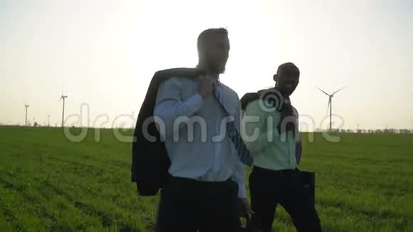 身穿商务服的黑人和白人正在绿色草地上行走背景是可见的风力发电机视频的预览图