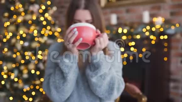 穿着毛衣的幸福微笑的女人在客厅的圣诞树旁喝着热巧克力圣诞节视频的预览图