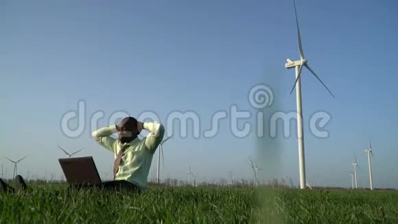 一个穿着西装的轻松的黑人坐在靠近风力发电机的绿色草地上视频的预览图