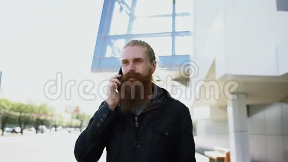 多莉拍摄的一个留胡子的年轻时髦男人集中在城市街上的电话上在办公楼附近交谈视频的预览图