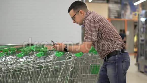 一个年轻人在他的智能手机屏幕上打印一条信息靠在超市的购物车上使用视频的预览图