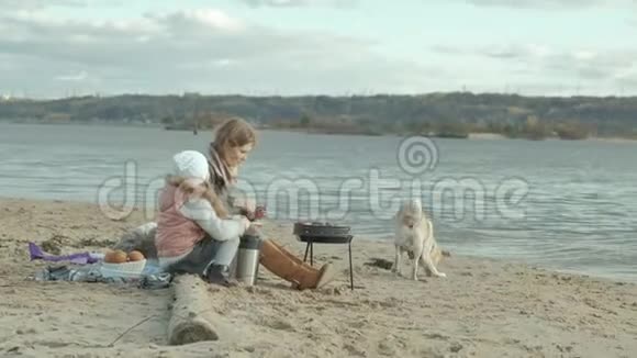 年轻的女人穿着外套有一个卷发的女孩母亲带着一个女儿坐在河边的海滩上大海有一个卷发视频的预览图