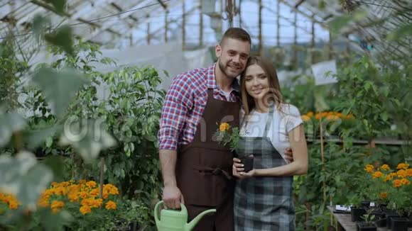 幸福的年轻夫妇在温室里微笑漂亮的女人和男人穿着围裙在花园里工作看着摄像机视频的预览图