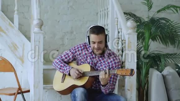 快乐迷人的男人坐在楼梯上带着无线耳机在家玩声吉他视频的预览图