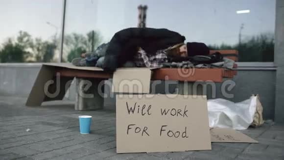迷路的无家可归的年轻人带着卡巴德标志睡在城市街的长凳上视频的预览图
