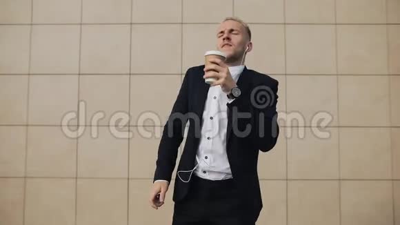 快乐的年轻商人带着咖啡在智能手机上用耳机听音乐在办公室附近跳舞视频的预览图