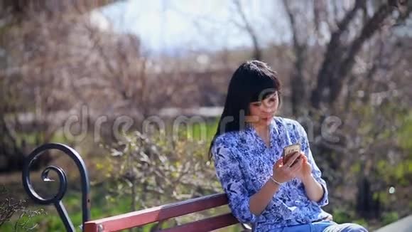一个美丽的亚洲女孩坐在公园里的长凳上在智能手机上读着一条信息非常惊讶哇哇大叫视频的预览图