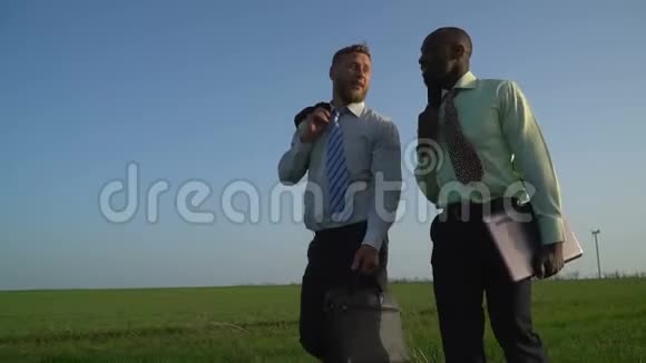 穿着商务服的黑人和白人正沿着绿色的草地行走远处可以看到风力发电机视频的预览图