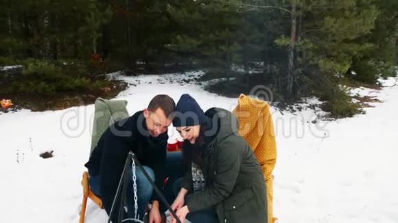 这对夫妇坐在冬天森林里的篝火旁坐在舒适的沙发上下雪了冬季快乐概念视频的预览图