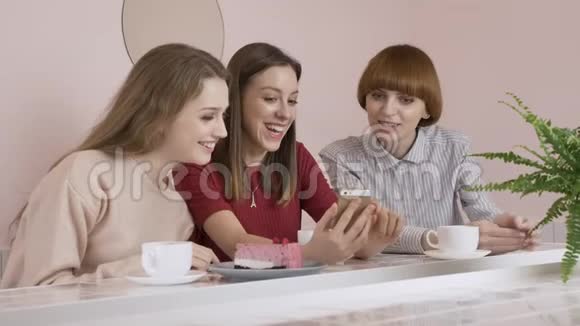 三个白种人年轻的女孩坐在咖啡馆里喝着咖啡吃着蛋糕微笑着笑着手势视频的预览图