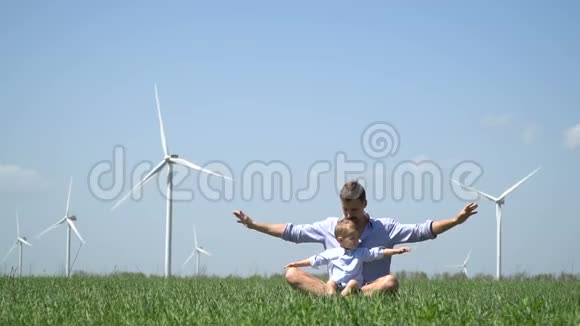 爸爸和儿子正坐在草地上双臂伸到风力发电机旁视频的预览图