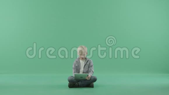 一个男孩带着他的平板电脑坐在绿色背景的地板上视频的预览图