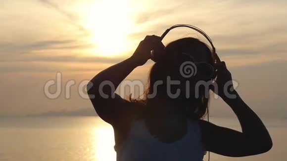 年轻的黑发快乐的女人带着耳机在美丽的日落海滩上听音乐跳舞慢慢慢慢视频的预览图