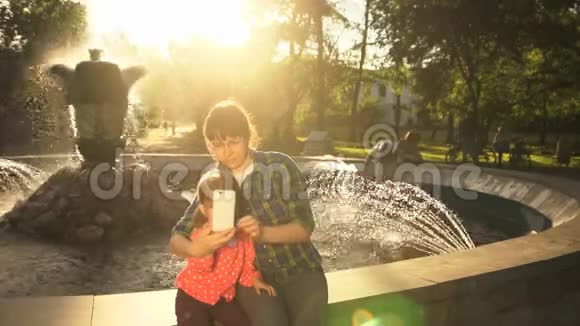 妈妈和女儿在日落时分在喷泉边打电话视频的预览图