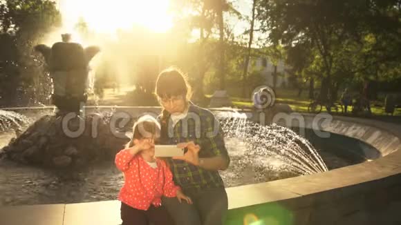 妈妈和女儿在日落时分在喷泉边打电话视频的预览图