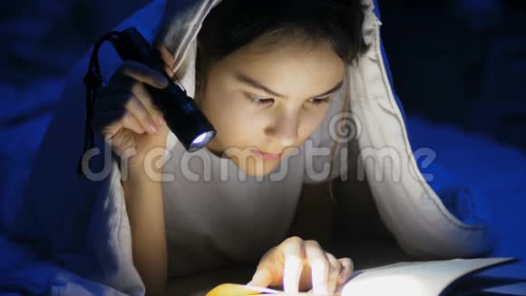 青少年女孩用手电筒在毯子下看书的特写镜头视频的预览图