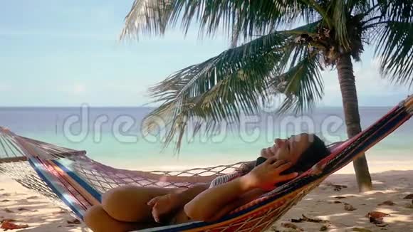 迷人的快乐女人在棕榈树旁边的热带海滩上在手机上聊天在吊床上放松美丽视频的预览图