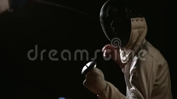 特写一个人戴着一个头盔用剑击击剑在黑暗的背景上与对手搏斗运动视频的预览图