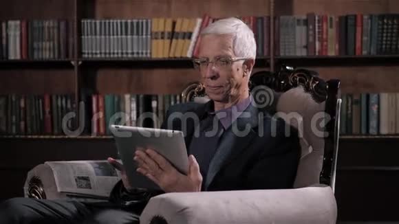 在家图书馆寻找平板电脑信息的老人戴眼镜和现代科技的快乐老人视频的预览图