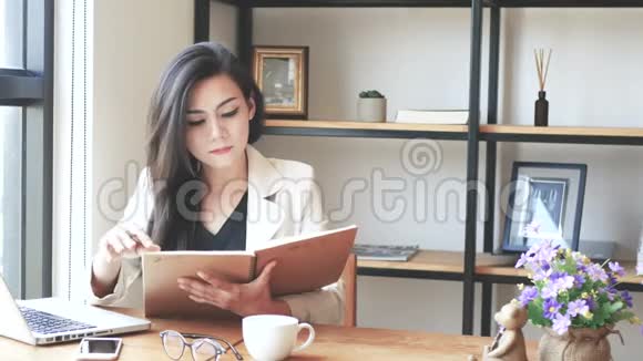 4K镜头美丽的亚洲商务女性坐着电脑手提电脑看书在咖啡馆里翻着便签页微笑视频的预览图