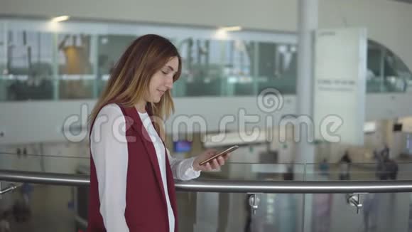 穿着优雅衣服的年轻女孩站在机场的智能手机里打字视频的预览图