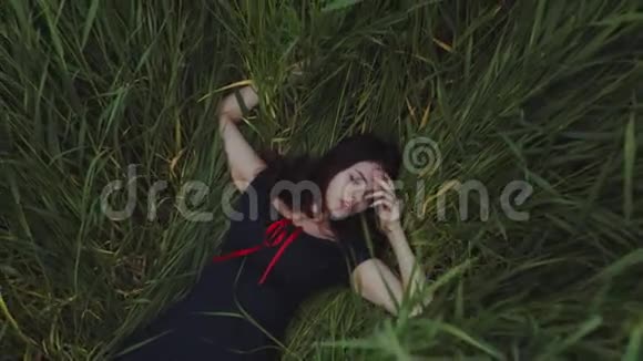 美丽的长发黑发女孩躺在绿色的高草抚摸触摸花浪漫气氛视频的预览图