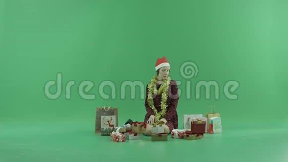 一个年轻的女人正坐在绿色的屏幕上打开她身边的空圣诞礼物视频的预览图