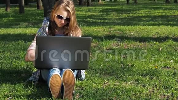 女孩在阳光明媚的一天在公园里带着笔记本电脑一个戴着太阳镜的少女一个十几岁的男孩坐在绿色的草地上视频的预览图